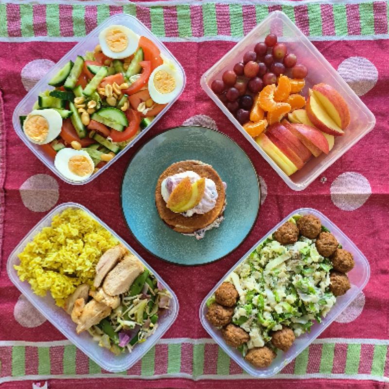 Meal prep op een roze kleed: salade met ei, fruit, pannenkoek en rijst met kip