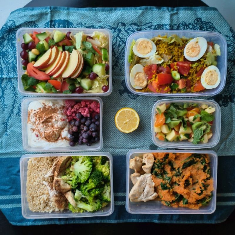 Meal prep op een groen kleed: appel, salades, kwark met fruit en zoete aardappel met kip