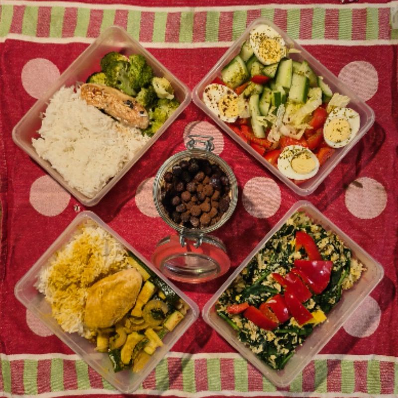 Meal prep op een rood kleed: rijst met kip, salades en groenten in bakjes