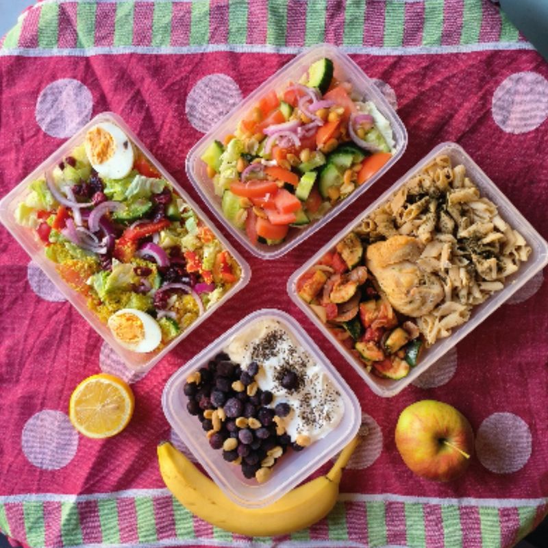 Meal prep op een roze kleed: salades, kip, kwark met noten en een banaan